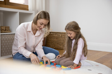 Young blonde mother and little beautiful daughter playing together indoor, sit on warm floor in modern nursery use wooden bricks enjoy playtime and communication spend time on weekend leisure at home
