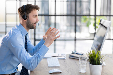 Cheerful young support phone male operator in headset, at workplace while using computer