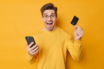 Cheerful handsome guy makes safe mobile banking via cellular application holds credit card provides...