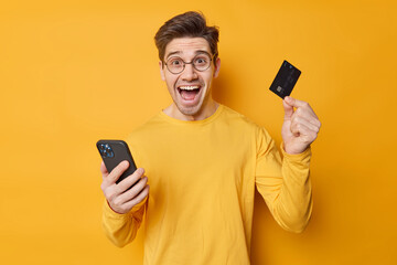 Cheerful handsome guy makes safe mobile banking via cellular application holds credit card provides internet payment wears round spectacles and casual jumper isolated over yellow background.