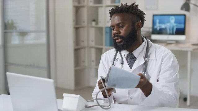 Modern young adult African American doctor wearing white coat sitting at desk having video call with patient
