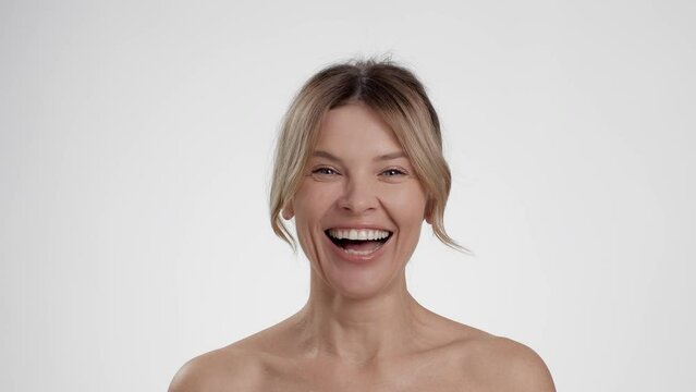 Happy aging. Carefree middle aged woman with bare shoulders laughing to camera, white studio background, slow motion
