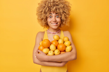 Surprised cheerful woman bites lips carries big heap of fresh oranges and lemons eats fruits full of vitamins dressed in casual t shirt isolated over vivid yellow background. People summer time - obrazy, fototapety, plakaty