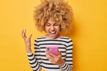 Angry outraged young woman with curly hair screams and focused at smartphone communicates in social networks wears casual striped jumper isolated over vivid yellow background. Negative emotions