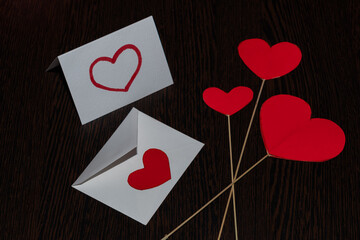 Red hearts made of cardboard, on straw stands. Red hearts and a white valentine card, with a white envelope, on a dark substrate with a woody texture.