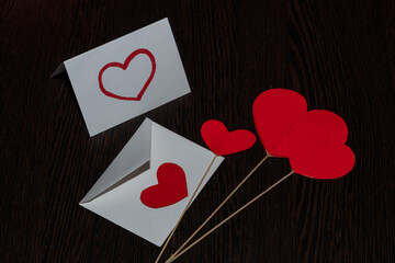 Red hearts made of cardboard, on straw stands. Red hearts and a white valentine card, with a white envelope, on a dark substrate with a woody texture.