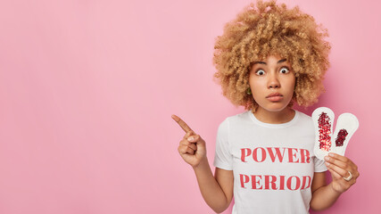 Horizontal shot of impressed woman with curly hair holds sanitary napkins uses hygienic products...