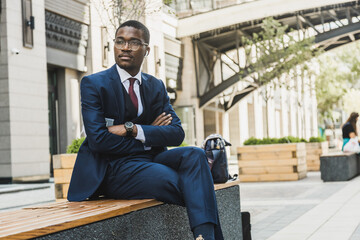 Portrait of a black African American businessman in a suit, glasses and headphones sits on a bench...