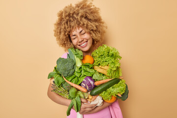Pleased smiling young woman with curly hair keeps eyes closed poses with harvested crops carries variety of green vegetables keeps to diet isolated over beige studio background. Spring vitamins
