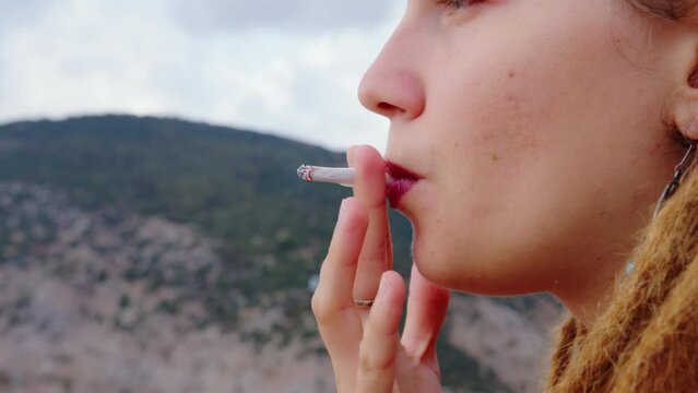 Girl tourist with dreadlocks smokes. Close up portrait. Thoughtful female traveler takes puff on cigarette