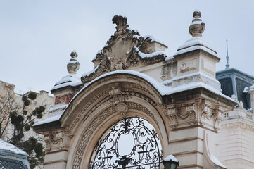 A snow-covered fragment of an ancient arch of gate with stucco elements, a curlicue in the center and a pommel with urns on the sides. Neoclassical architecture in Lviv, Ukraine.