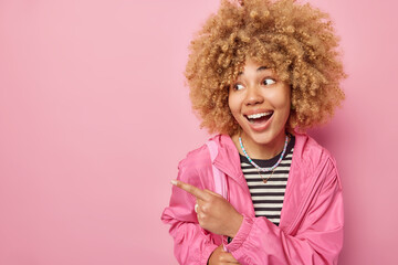 Cheerful impressed young woman with curly hair points away on blank space demonstrates awesome advertisement wears striped jumper and jacket isolated over pink background. Wow just look there