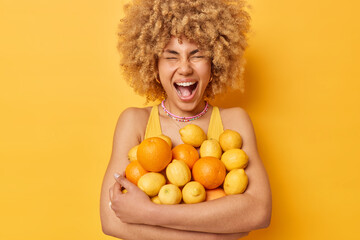 Emotional cheerful woman embraces heap of fresh oranges and lemons eats tropical fruits full of...