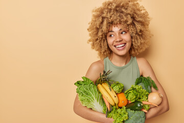Horizontal shot of beautiful woman carries variety of fresh vegetables and fruits consumes grocery full of vitamins looks away stands against beige background blank space for your promotional content