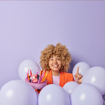 Vertical Shot Of Cheerful Young Woman With Curly Hair Points Index Finger Above Shows Something Poses Around Inflated Helium Balloons Holds Delicious Festive Cake Celebrates Birthday Anniversary