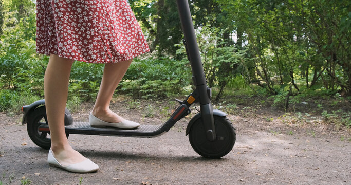 Woman in dress rides on an electric scooter along a path in a park with green trees. Eco concept, green energy, vehicle. Side view, daytime, spring summer. - Powered by Adobe