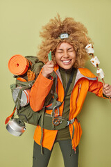 Cheerful female camper holds roasted marshmallow has picnic during spare time wears orange jacket returns home from hiking trip carries rucksack isolated over dark green background. Adventure tourism