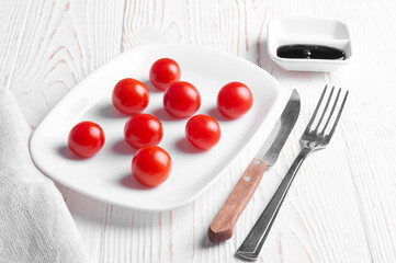 cherry tomatoes lie on a white plate on a white wooden background, a knife and a fork lie nearby and there is a balsamic sauce