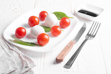 caprese salad of cherry tomatoes, mozzarella cheese balls and spinach leaves lie in a white plate, knife and fork, and balsamic sauce on a white wooden background