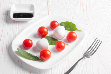 Naklejka na ściany i meble caprese salad of cherry tomatoes, mozzarella cheese balls and spinach leaves and balsamic sauce lie in a white plate, and a fork lies next to it on a white wooden background