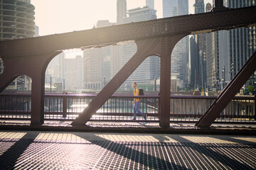 Side view of man on the move while walking on bridge and looking around. City in morning light. . - obrazy, fototapety, plakaty