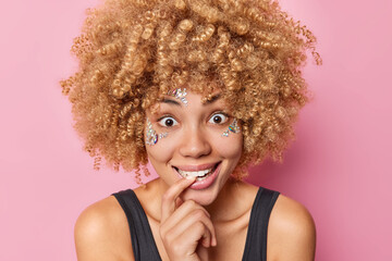 Headshot of happy surprised young woman bites finger looks wondered has face decorated with shiny stones has natural curly hair dressed in casual black t shirt isolated over pink background.