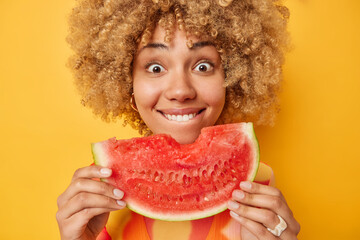 Close up shot of curly haired positive woman holds big slice of juicy watermelon enjoys eating her...