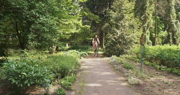 young woman rides a folding bike through a park with green trees towards the camera. The concept of a healthy lifestyle, eco movement, green transport. Wide shot, spring summer, daytime