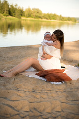 mom with a newborn baby sits on the beach, on the sand. mom holds a newborn baby in her arms.