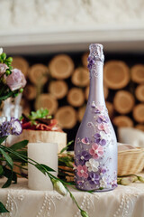 Decorative purple bottle of champagne, with patterns of flowers, candles and flowers, at the festive table for the newlyweds in a cozy restaurant by the fireplace with firewood.