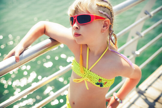 A little girl in a yellow swimsuit, red glasses and with a fashionable hairstyle, stands at the railing next to the sea, on a bright sunny day.