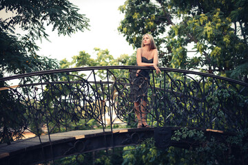 Naklejka na ściany i meble A slender elegant girl in a dark dress with blond hair stands in a park alley on a bridge against the backdrop of trees in summer.