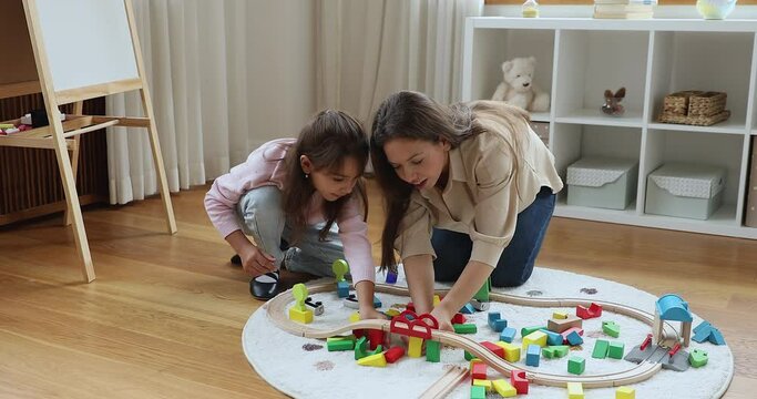 Woman her daughter play wooden cubes at home. Loving mother and little cute girl seated on warm floor in nursery enjoy playtime with railroad and colored bricks. Child games with modern toys concept