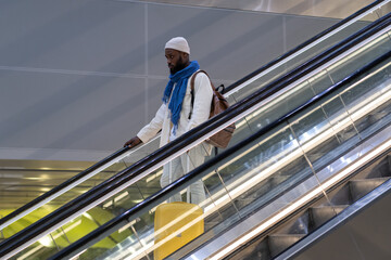 Young African American passenger man with yellow luggage stands on escalator, arrives from abroad, holds handrail in airport terminal. Trendy Black traveler man with suitcase at the station.