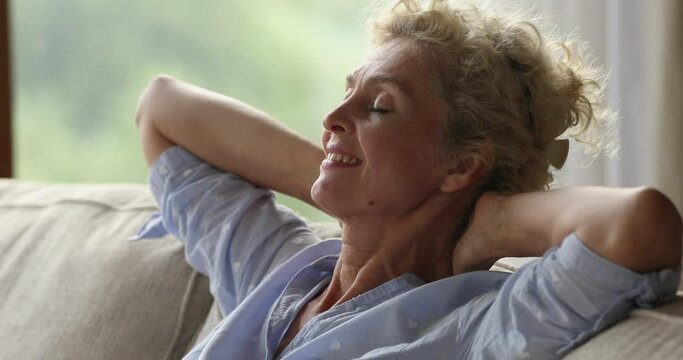Close up serene older 60s dreamy female resting leaned on soft sofa cushions feel good, breath fresh air enjoy relaxation at modern cozy home with climate control. Vacation and accommodation concept