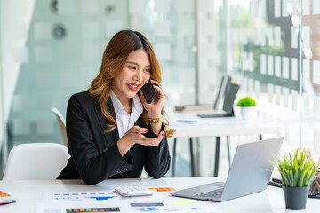 Happy business asian manageress working at her desk in the office taking a call on her mobile phone while writing notes on a notepad