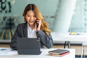 Happy business asian manageress working at her desk in the office taking a call on her mobile phone while writing notes on a notepad