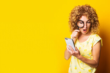 surprised curly young woman with a magnifying glass looks into the phone on a yellow background.