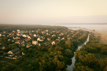 Residential private houses on the banks of the river among the trees on a foggy summer morning - aerial drone shot. Foggy morning in the ecovillage: houses near the water among dense green trees.