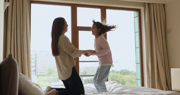 Active adorable little girl jumping on bed in modern bedroom while holding mommy hands, feeling carefree, having fun together at home. Playtime and leisure, happy holidays and accommodation concept