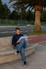 A girl in jeans and a blue jacket sits on a stone bench in the park and reads documents. A foot on the other leg. In the background there is a palm tree, a red carpet for running and a roadway. The