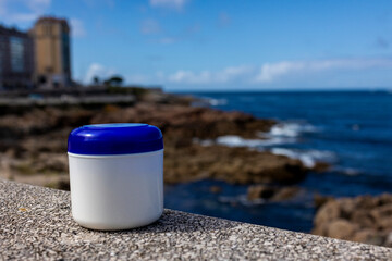 A white cream jar with a blue lid stands on a stone surface. There is a blue sky in the background. Blue okeean, stone islet shore and urban environment. Close-up.