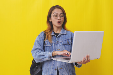 Surprised young asian college student standing while holding laptop. Isolated on yellow background