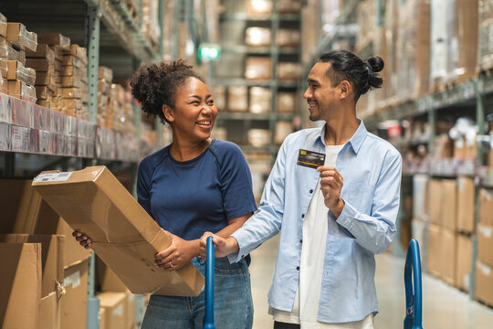 African woman picks up a box of furniture she has bought and an Asian man, her boyfriend holding a credit card, is comfortable to use in a wholesale store warehouse.