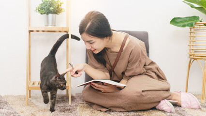 Pet lover concept, Young Asian woman playing with cat and taking notes on notebook in living room