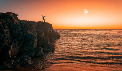 silhouette of saxophonist playing on cliff at sunset and full moon in the background..