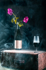 Bodegón de botella de vino tinto encima de una maleta vintage con rosas rosas y humo, alto contraste, fondo negro