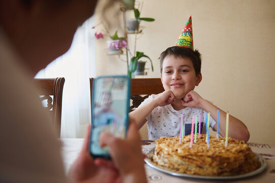 Cheerful Caucasian teenage boy sitting at table with birthday cake with candles smiling while posing to his blurred mom taking photo of him on smartphone in foreground. Birthday party concept