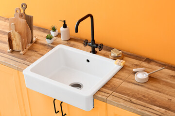 Fototapeta na wymiar Wooden counter with ceramic sink and cleaning supplies near orange wall in kitchen