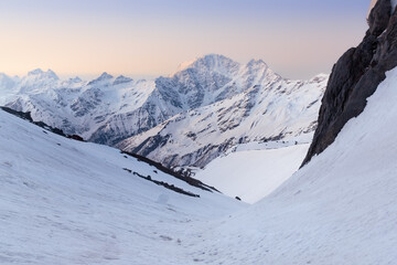 Fototapeta na wymiar Snow-covered mountain peaks in pastel sunrise colors with dark rocks in foreground, Caucasus, Russia